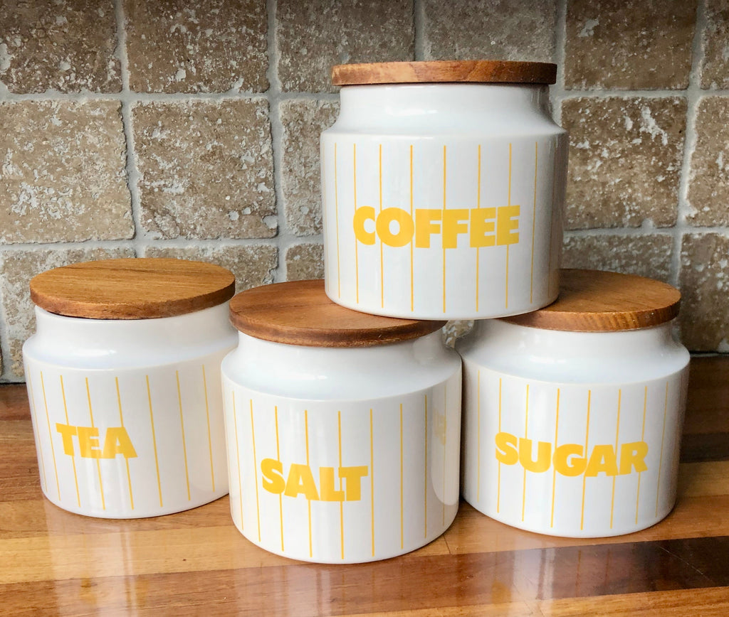Four Hornsea pottery Stripes ceramic canisters with wooden lids for coffee, tea, salt, and sugar, decorated with yellow vertical stripes and bold yellow text, arranged on a wooden surface against a tiled wall.