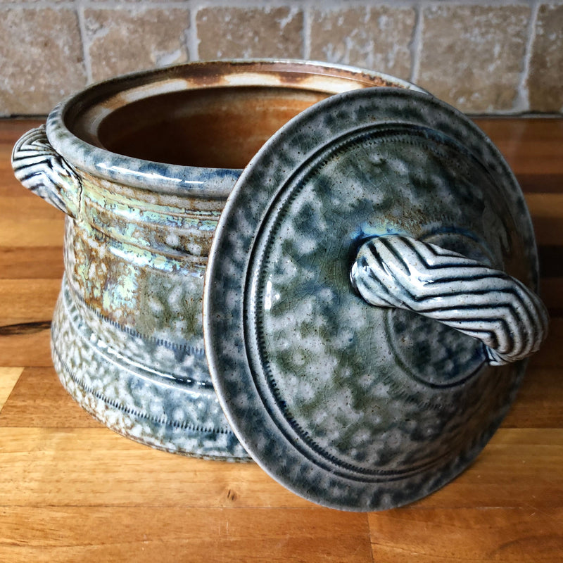 Lid leaning beside pot: Jane Hamlyn stoneware casserole with lid leaning against the pot, showing its blue-grey glaze and chevron lid handle.
