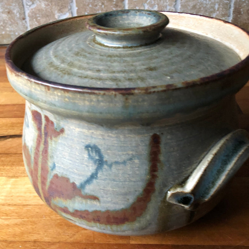 Angled view of Vera Tollow casserole showing free-flowing rust-red and blue brushwork beside the grooved side handle.